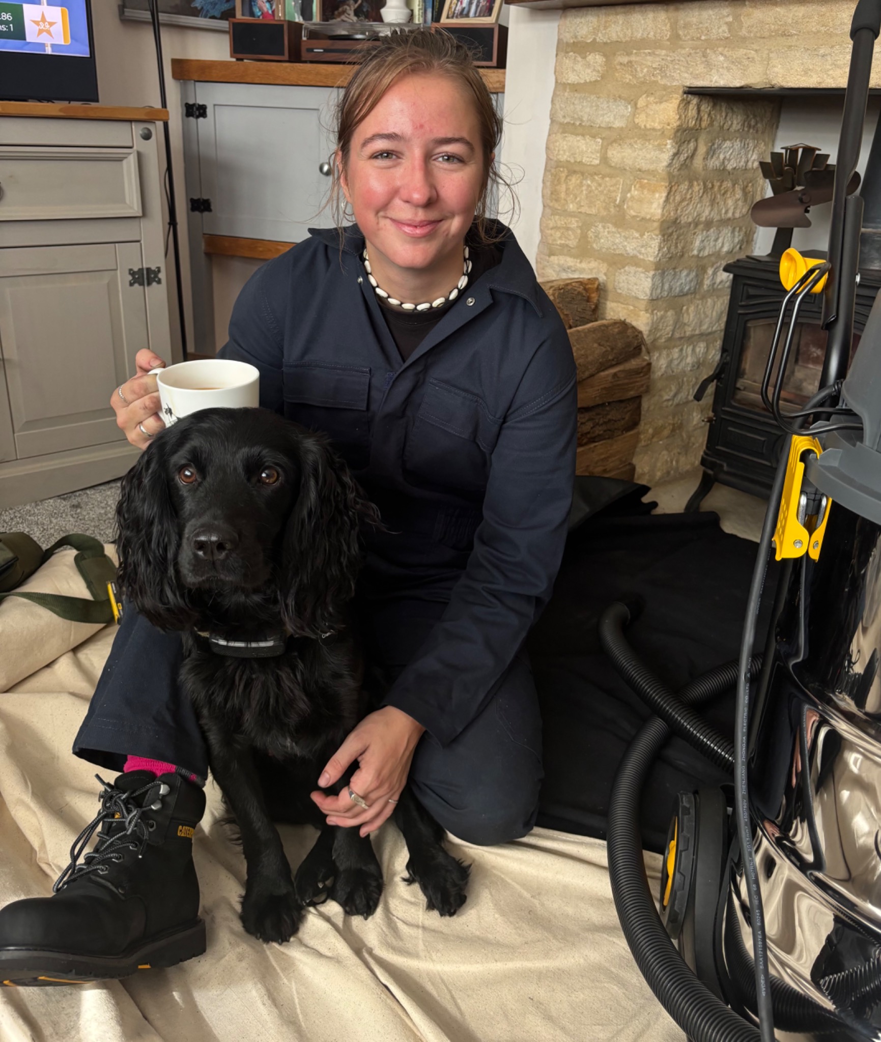 Megan enjoying a cup of tea with a black spaniel beside her sweeping equipment.