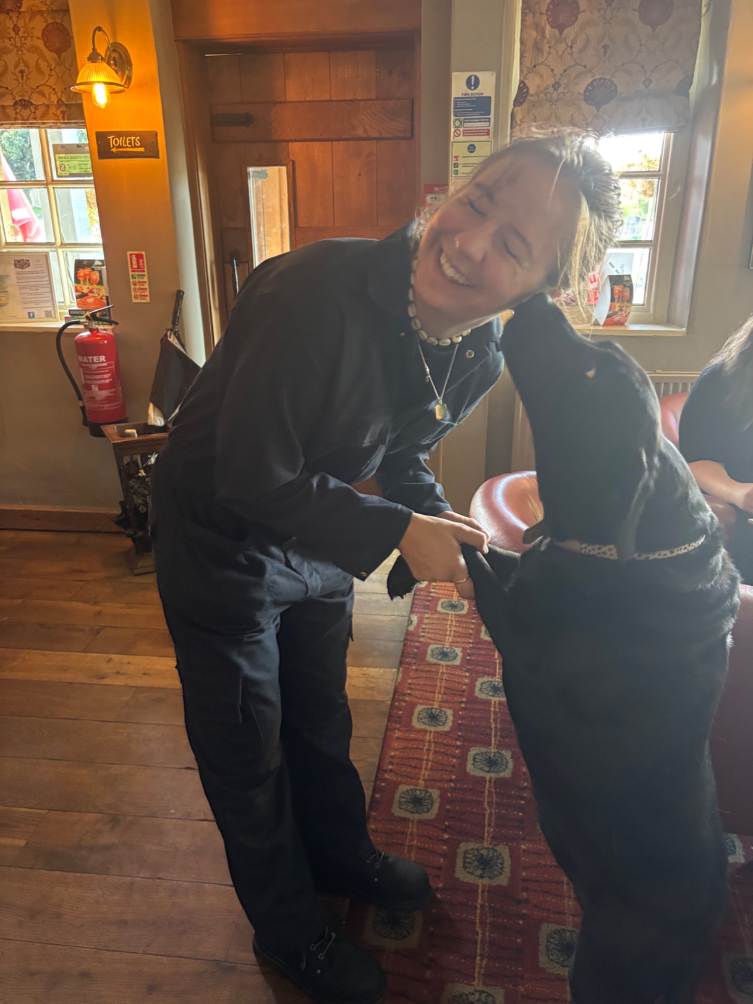 Megan smiling and holding paws with a friendly black dog inside a pub.