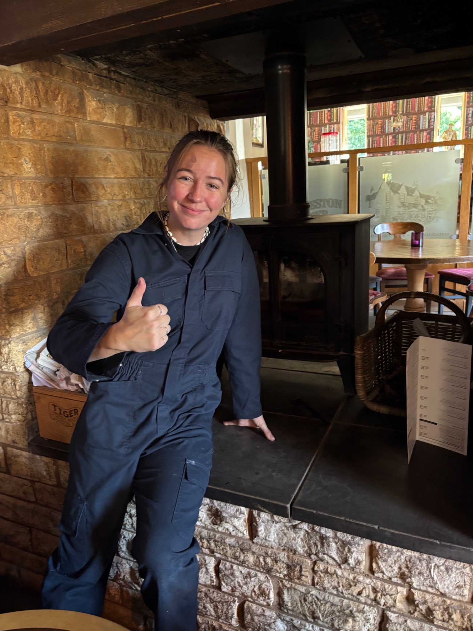 Megan giving a thumbs up beside a log burner in a stone fireplace.